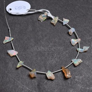 Peut inclure: Un collier de pierres précieuses d'opale brute, chacune avec des formes uniques et des couleurs irisées orange, verte et blanche. Les pierres sont enfilées sur un fil fin de couleur argentée. Une étiquette blanche avec un numéro manuscrit est attachée.