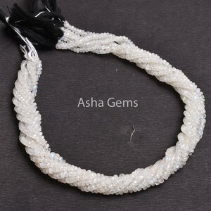 May include: A strand of white moonstone beads, approximately 15 inches long, with a black tassel at the end. The beads are faceted and have a subtle iridescent sheen.  Aisha Gems is printed on the background.