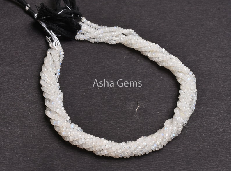 May include: A strand of white moonstone beads, approximately 15 inches long, with a black tassel at the end. The beads are faceted and have a subtle iridescent sheen.  Aisha Gems is printed on the background.