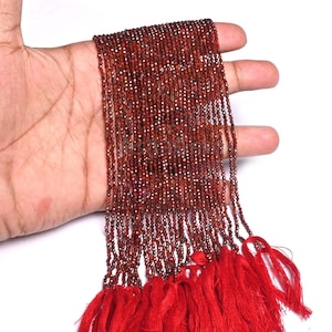 Może przedstawiać: Wiele sznurów małych, ciemnoczerwonych koralików, prawdopodobnie granatów, trzymanych w dłoni. Koraliki są nawleczone, z czerwoną frędzlą na dole. Koraliki mają średnicę około 2 mm.