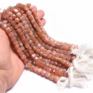 May include: A strand of peach-colored sunstone beads, each bead is faceted and has a square shape. The beads are strung on a white cord with white tassels at the ends.