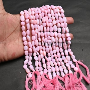 Puede incluir: Se muestran hebras de cuentas rosas ovaladas, probablemente piedras preciosas. Las cuentas son de un color rosa claro y suave y parecen estar ensartadas. Se ven borlas rosas en la parte inferior de las hebras. Las cuentas se sostienen en una mano.