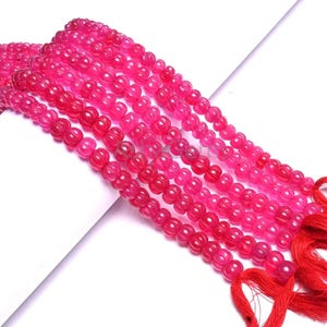 May include: Strands of vibrant pink beads with a glossy finish. The beads are strung together, forming multiple lines. Red tassels are attached to the ends of the strands. The beads are likely for jewelry making.