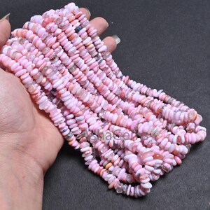 Puede incluir: Una hebra de cuentas de ópalo rosa, con una variedad de formas y tamaños, se muestra en una mano. Las cuentas son de color rosa pálido con algunos acentos blancos y naranja.