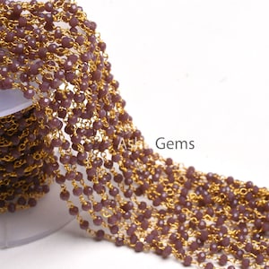 May include: Close-up of a beaded chain with small, faceted, purple beads strung on gold-colored chain links. The chain is partially rolled on a white spool, with the text "Ash Gems" visible in the lower right corner.