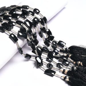 Könnte beinhalten: Eine Strähne aus schwarzen facettierten Edelsteinperlen mit silbernem Draht, der durch die Mitte jeder Perle gezogen ist. Die Perlen sind rechteckig und haben eine glänzende, polierte Oberfläche. Die Strähne ist mit einem schwarzen Quastenbesatz versehen.