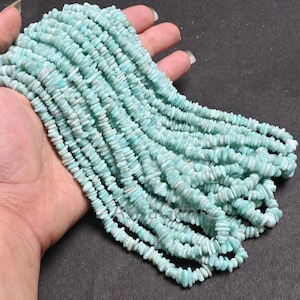 Puede incluir: Un largo hilo de pequeñas piezas irregulares de piedra amazonita en tonos azul verdoso claro. Las cuentas están ensartadas en una línea continua, mostrando una textura natural mate y formas variadas características de fragmentos de piedra pulida.