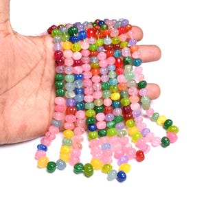 Puede incluir: Cadenas de cuentas coloridas en varios tonos de rosa, verde, amarillo, azul y rojo. Las cuentas son pequeñas y redondas, con una superficie texturizada. Las cuentas se sostienen en una mano sobre un fondo blanco. Ideales para la creación de joyas.