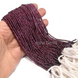 Könnte beinhalten: Eine Strähne kleiner, facettierter Rubinperlen in tiefroter Farbe. Die Perlen sind auf einem weißen Faden aufgereiht und haben einen Durchmesser von etwa 2 mm. Die Perlen sind in einer Hand zu sehen, wobei die Strähne nach rechts verläuft.