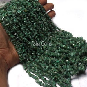 Puede incluir: Una hebra de cuentas de chip de color verde esmeralda. Las cuentas son de forma irregular y tienen una textura natural y rugosa. Las cuentas están enhebradas en un hilo transparente y tienen aproximadamente 10 mm de largo.