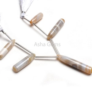 May include: Six elongated, polished gemstone beads with a light brown and white striped pattern. The beads are strung on silver-colored pins, with white tassels visible in the background. The beads are likely for jewelry making.
