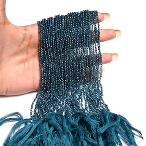 Puede incluir: Varias hebras de pequeñas cuentas de color verde azulado se sostienen en una mano. Las cuentas están ensartadas, creando un efecto fluido. La parte inferior de las hebras están atadas con hilos de color verde azulado. Las cuentas tienen un ligero brillo.