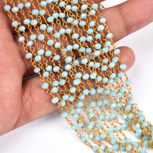 Puede incluir: Una hebra de cuentas facetadas de color azul claro enhebradas en una cadena dorada. Las cuentas son pequeñas y de tamaño uniforme. La cadena es delicada y tiene un brillo sutil.