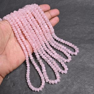 Puede incluir: Varias hebras de cuentas de cuarzo rosa rosa claro. Las cuentas son pequeñas, redondas y de tamaño uniforme. Las hebras se sostienen en una mano sobre un fondo gris oscuro. Las cuentas tienen un aspecto suave y translúcido.