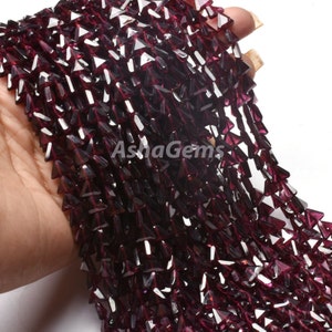 May include: A strand of deep red garnet gemstone beads, each bead is faceted into a triangular shape. The beads are strung together to create a long, flowing strand.