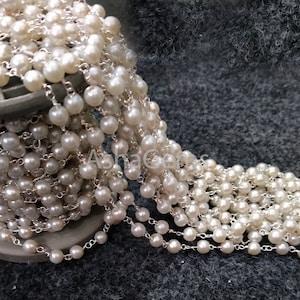 May include: Strand of white, round pearls strung on a silver chain. The pearls vary slightly in size and have a lustrous sheen. The chain is tightly woven, creating a delicate yet durable design. The pearls are displayed on a gray surface.