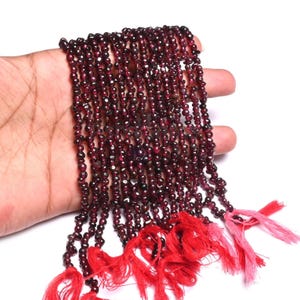 Könnte beinhalten: Mehrere Stränge kleiner, dunkelroter Granatperlen. Die Perlen sind aufgefädelt, mit roten und rosa Quasten unten. Die Perlen werden vor einem weißen Hintergrund präsentiert.