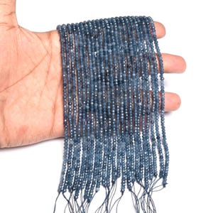 Könnte beinhalten: Mehrere Stränge kleiner, facettierter, blau-grauer Perlen. Die Perlen sind auf schwarzem Faden aufgereiht und werden in einer Hand gehalten. Die Perlen haben einen subtilen Glanz, wahrscheinlich für die Schmuckherstellung. Jeder Strang ist etwa 40 cm lang.