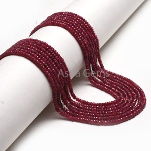 May include: A strand of deep red ruby beads, faceted and polished, arranged in a loose coil on a white surface. The beads are uniform in size and shape, creating a consistent and elegant look.