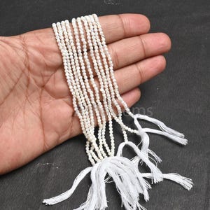 Puede incluir: Múltiples hebras de pequeñas cuentas blancas y redondas con un brillo nacarado. Las cuentas están ensartadas, con borlas blancas que aseguran las hebras en la parte inferior. Las cuentas se sostienen en una mano sobre un fondo oscuro.