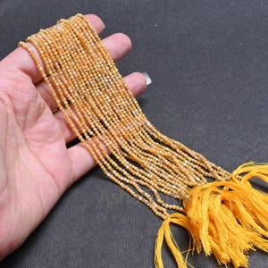 Puede incluir: Múltiples hebras de pequeñas cuentas redondas de piedras preciosas de color amarillo anaranjado. Las cuentas se sostienen en una mano, con varias hebras visibles. Borlas amarillas brillantes están unidas a los extremos de las hebras. El fondo es gris oscuro.