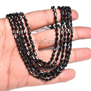Peut inclure: Présentation de cinq rangées de perles de pierres précieuses noires. Chaque rangée est composée de petites perles en forme de goutte. Les perles ont une finition brillante et sont enfilées ensemble, créant un effet superposé. Les perles sont tenues dans une main.