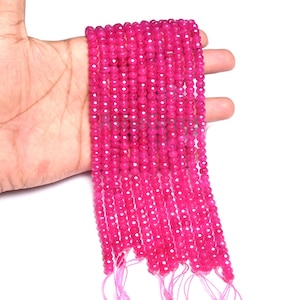 Puede incluir: Cadenas de cuentas facetadas de piedras preciosas de color rosa intenso. Las pequeñas cuentas redondas están ensartadas, creando una apariencia texturizada. Las cuentas se sostienen en una mano sobre un fondo blanco.