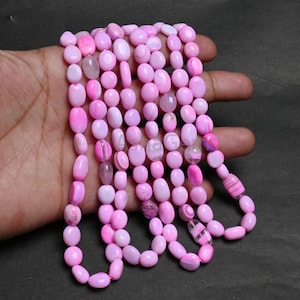 Puede incluir: Un collar de cuentas rosas ovaladas en diferentes tonos de rosa y blanco. Las cuentas están ensartadas, creando un collar o una pulsera. Las cuentas son lisas y pulidas, reflejando la luz.