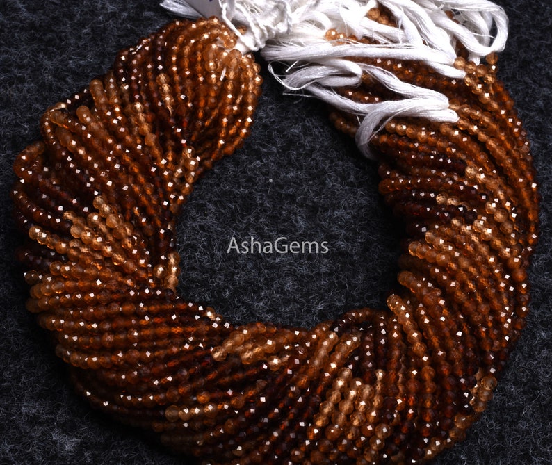 May include: A strand of brown gemstone beads, likely citrine, with a faceted finish. The beads are strung on a white thread and are arranged in a circular shape. The image includes the text "AshaGems".