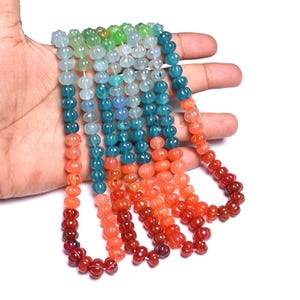 Puede incluir: Hilos de cuentas de piedras preciosas de colores en tonos verdes, azules, naranjas y rojos. Las cuentas tienen una forma texturizada única. Las cuentas se sostienen en una mano sobre un fondo blanco.