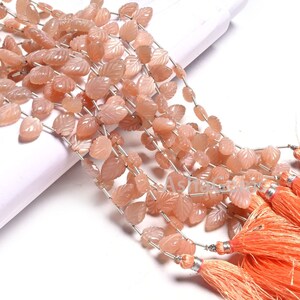 Peut inclure: Un brin de perles de pierre de soleil de couleur pêche, sculptées en forme de feuilles, avec un fil d'argent entre chaque perle. Les perles sont enfilées sur un fil d'argent et ont un pompon à l'extrémité.