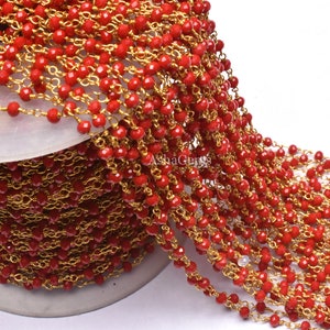 Puede incluir: Un carrete de cuentas de vidrio facetadas rojas enhebradas en una cadena dorada. La cadena está hecha de pequeños anillos unidos. Las cuentas son pequeñas y redondas, y están espaciadas uniformemente a lo largo de la cadena.