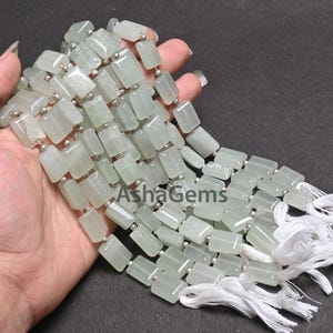 Puede incluir: Una hebra de cuentas de piedras preciosas verde claro, cada cuenta es rectangular y tiene un acabado liso y pulido. Las cuentas están enhebradas en alambre plateado y tienen borlas blancas en los extremos.