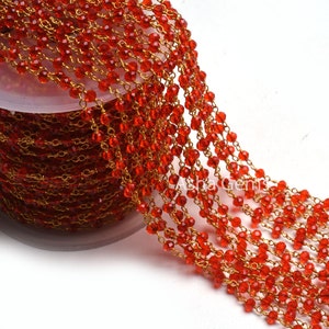 May include: Close-up of a spool of red beaded chain with gold-colored wire. The beads are small, faceted, and a vibrant red color. The chain is wound around a white spool, and the beads are strung together with gold-colored wire.