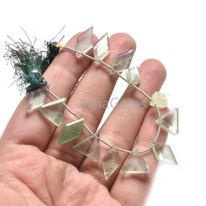 Puede incluir: Un hilo de cuentas de piedras preciosas de color verde pálido facetadas en varias formas geométricas, incluyendo diamantes y triángulos, ensartadas en un alambre plateado fino con una borla verde oscuro en un extremo. Las cuentas son translúcidas.