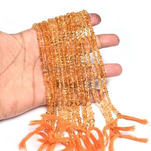 以下が含まれることがあります： 小さなオレンジイエローの宝石ビーズのストランド。ビーズは手で持たれており、複数のストランドが見えます。ストランドの端はオレンジ色のタッセルで結ばれています。ビーズはファセット加工された外観と半透明の品質を持っています。