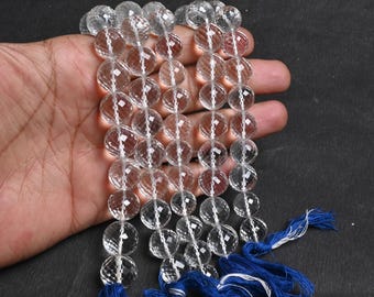May include: Strands of clear, faceted quartz beads. Each bead is round and has multiple facets that catch the light. The strands are held together with white string and finished with blue tassels. The beads are displayed against a dark background.