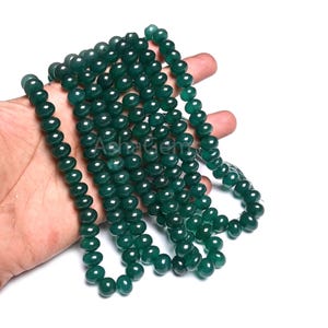 Puede incluir: Un collar de cuentas redondas de color verde oscuro. Las cuentas están ensartadas, formando un collar o una pulsera. Las cuentas se sostienen en una mano sobre un fondo blanco. Las cuentas parecen pulidas y tienen un acabado brillante.