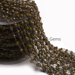 Puede incluir: Cadena de cuentas verde oliva con eslabones de metal gris oscuro. Las cuentas, pequeñas y facetadas, están muy juntas. La cadena está enrollada, lo que sugiere que se vende por metros para la fabricación de joyas u otras manualidades.