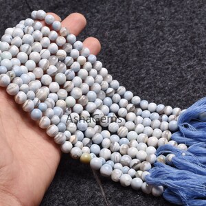 Puede incluir: Una hebra de cuentas de ágata azul claro y blanco con un patrón rayado. Las cuentas son redondas y tienen un acabado liso y pulido. La hebra mide aproximadamente 45 cm de largo y está acabada con borlas azules.
