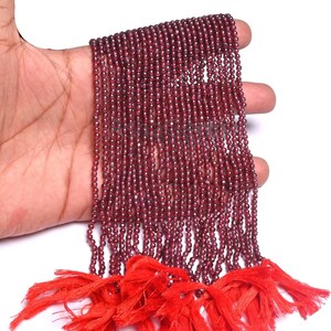 Puede incluir: Hebras de pequeñas cuentas redondas de color rojo intenso, posiblemente granate, se muestran sobre un fondo blanco. Las cuentas están ensartadas, con una borla roja brillante en la parte inferior. Las cuentas son de un color intenso y rico.