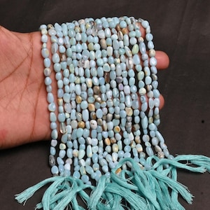 Puede incluir: Cadenas de cuentas de piedras preciosas de color azul claro, con una mezcla de tonos blancos y marrones. Las cuentas ovaladas están ensartadas, con una borla turquesa en la parte inferior. Las cuentas se sostienen en una mano.