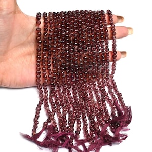 May include: Strands of small, round, faceted beads in a deep burgundy color. The beads are strung together, with tassels at the bottom. The beads are held in a hand against a white background.