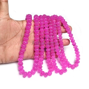 May include: A collection of bright pink beaded necklaces. The beads are round with a textured, floral pattern. The necklaces are held in a hand against a white backdrop. The beads are a vibrant, eye-catching colour, perfect for jewellery making.