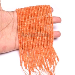 Può includere: Primo piano di fili di piccole perline rotonde arancioni. Le perline sono infilate insieme, creando una superficie strutturata. Le perline sono tenute in una mano su uno sfondo bianco. Le perline sono di un colore arancione vibrante e caldo.