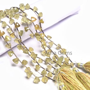 May include: A strand of light green gemstone beads with a triangular faceted shape. The beads are strung on a silver wire and have a metallic sheen. The strand is displayed on a white surface with two green tassels.