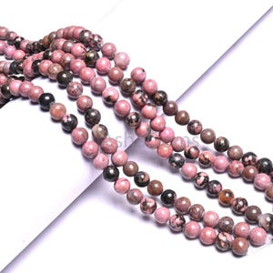 May include: Strands of pink and brown gemstone beads. The beads are round and vary in color, with some having black and white speckles. The beads are strung together, creating a necklace or bracelet.