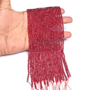 Puede incluir: Un hilo de pequeñas cuentas facetadas de color rojo rubí. Las cuentas están ensartadas en un hilo rojo, creando un efecto de cascada. Las cuentas se sostienen en una mano sobre un fondo blanco. Las cuentas parecen estar hechas de un material translúcido.