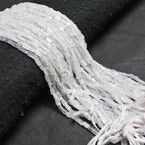 Puede incluir: Primer plano de una colección de cuentas blancas rectangulares. Las cuentas están ensartadas, creando un efecto de cascada. Las cuentas translúcidas reflejan la luz. Los hilos se juntan con hilos blancos, lo que sugiere un componente de joyería o un suministro artesanal.