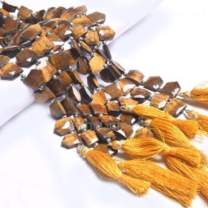 Könnte beinhalten: Eine Kette aus braunen und goldenen Tigerauge-Edelsteinen mit facettierten Kanten. Die Perlen sind auf einem Silberdraht aufgefädelt und haben eine polierte Oberfläche. Die Kette ist mit goldenen Quasten verziert.
